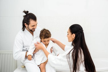 Parents smiling and looking at toddler son in white bathrobe  clipart