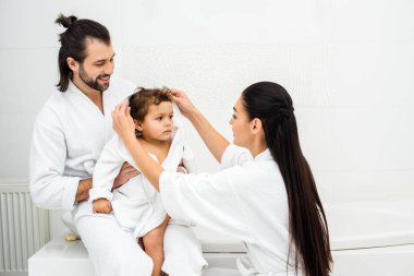 Parents sitting in bathroom with toddler son in white bathrobe  clipart
