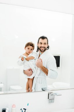 Dad and toddler son looking in mirror at home in white bathroom  clipart