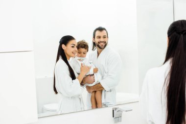 Mom and dad looking in mirror and holding toddler son in white bathrobes  clipart