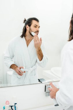 Handsome man in white bathrobe shaving in front of mirror clipart