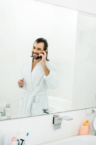 handsome adult man talking on smartphone and brushing teeth in bathroom