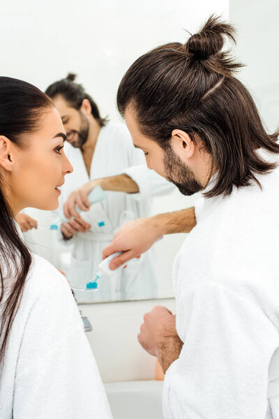 happy couple in white bathrobes brushing teeth together in bathroom