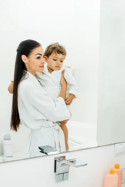 美丽的母亲抱着儿子在白色浴衣在浴室 — 图库照片