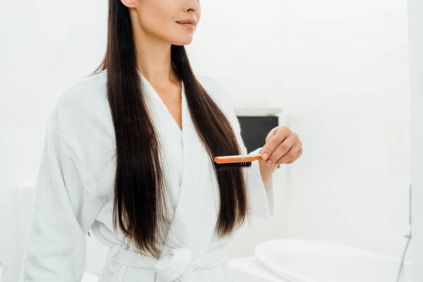 cropped view of woman combing long hair with hairbrush in bathroom