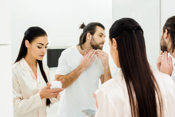 beautiful woman holding body cream while handsome man looking at mirror