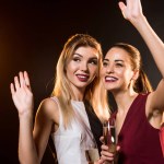 Belles jeunes femmes en robes avec des verres de champagne agitant sur noir lors de la fête du Nouvel An