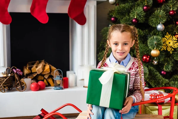 cute smiling child sitting on sled and holding christmas present