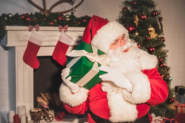 Weihnachtsmann Hält Und Schüttelt Weihnachtsgeschenk Während Kamin Sitzt — Stockfoto