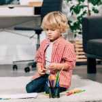 Lockige kleine Junge sitzt auf dem Boden mit bunten Filzstiften und weißem Papier zu Hause Lockige kleine Junge sitzt auf dem Boden mit bunten Filzstiften und weißem Papier zu Hause