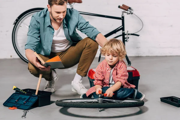 有选择的焦点人与书坐在小儿子附近修理自行车轮子用钳子在车间 — 图库照片