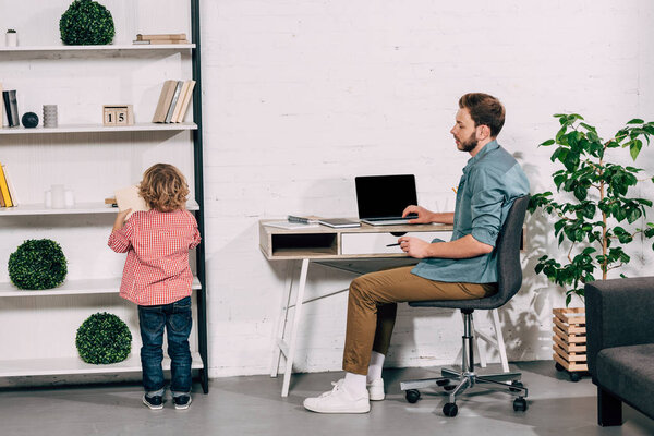 rear view of boy putting book on shelf while his father sitting behind at table with laptop 