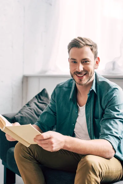 Laughing Young Man Holding Book Looking Camera Sofa Home — Free Stock Photo