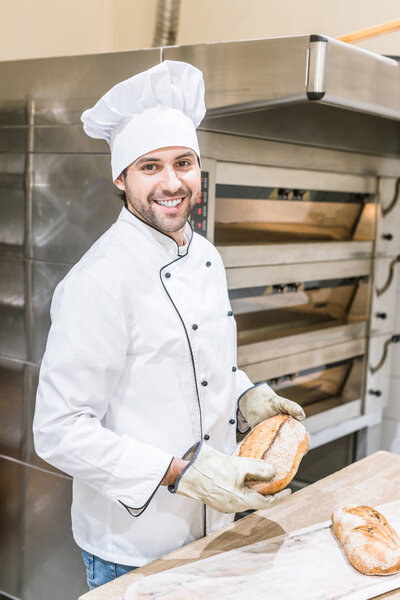smiling baker in chefs uniform holding fresh bread near oven
