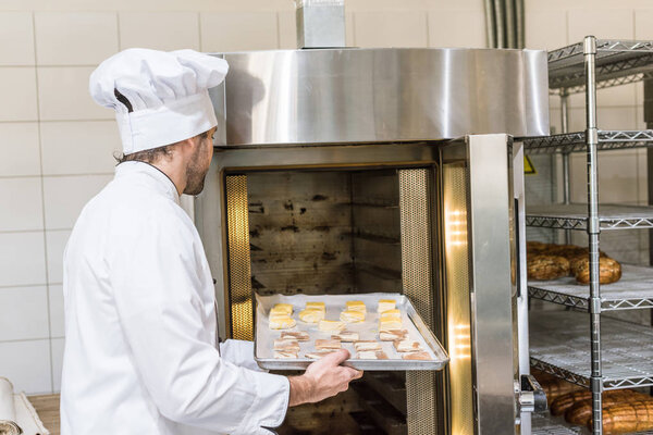 male baker in chefs uniform putting raw dough in oven