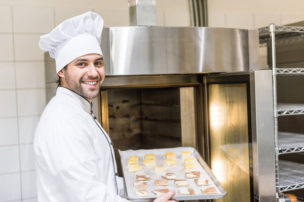 smiling baker in white chefs uniform putting baking tray with uncooked dough in oven