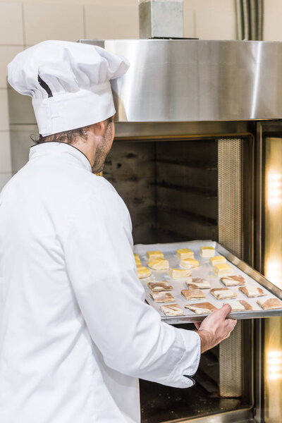male baker in white chefs uniform putting baking tray with uncooked dough in oven