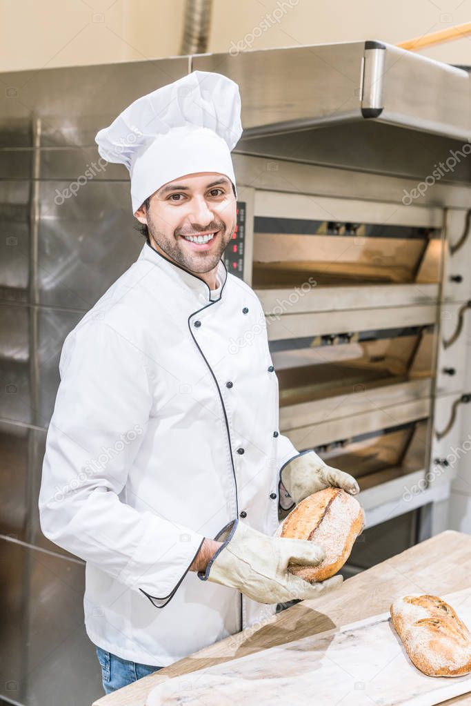 smiling baker in chefs uniform holding fresh bread near oven