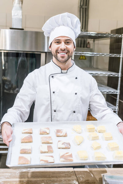 smiling baker in chefs uniform holding baking tray with raw dough pieces