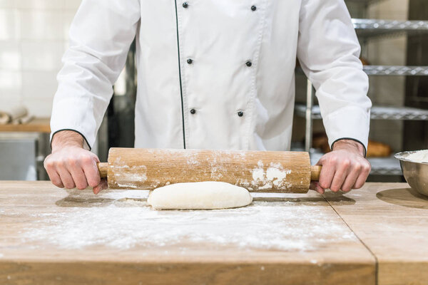close up of baker hands rolling out raw dough at wooden counter