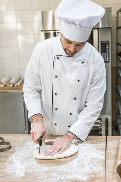 Baker in white chefs uniform cutting dough on wooden table at professional kitchen