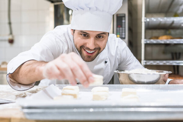 Handsome baker smiling and laying uncooked dough on tray