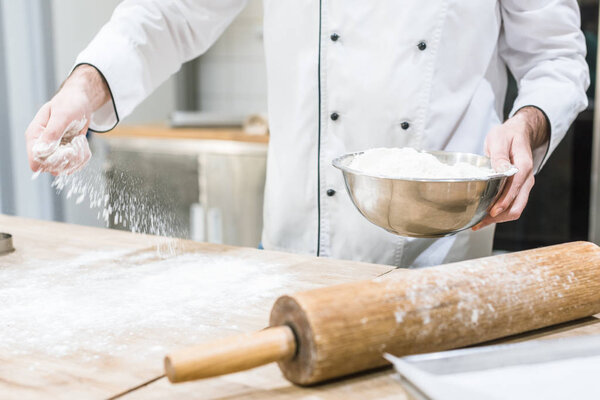 Cropped view of baker holding metal bowl and scattering flour on wooden table
