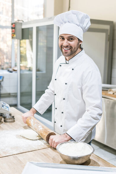 Handsome chef smiling and rolling out dough in professional kitchen