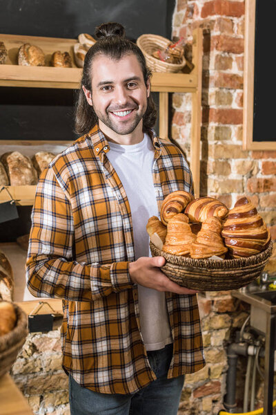 Smiling seller holding wicker basket with pastry in bakehouse