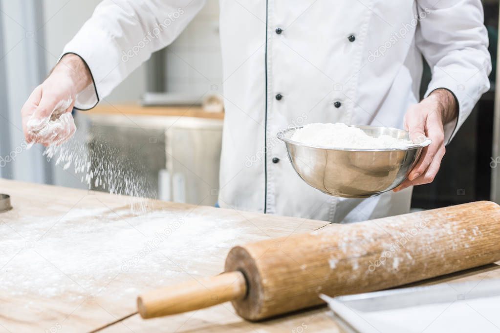 Cropped view of baker holding metal bowl and scattering flour on wooden table