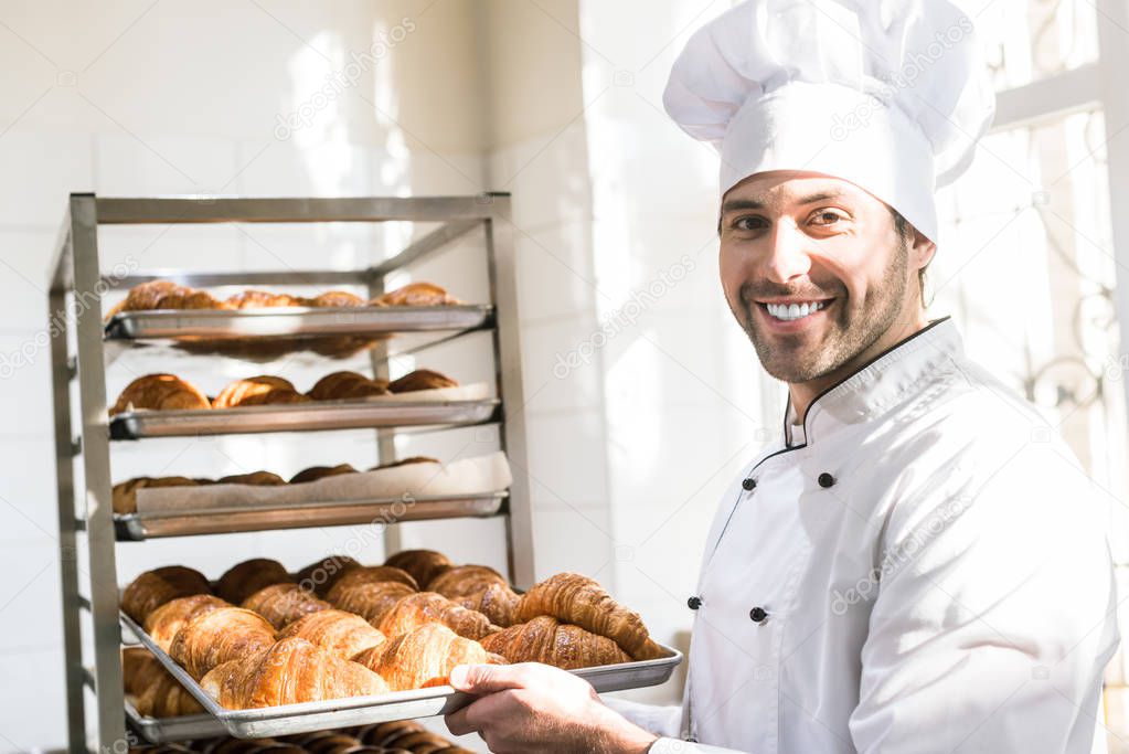 Smiling baker holding tray with fresh cooked croissants in bakehouse