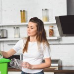 Woman putting plastic bottles in green recycle box at wooden table in kitchen