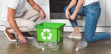 cropped view of couple standing at kitchen with green recycle box and plastic bottles on floor  clipart