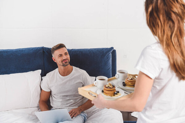 Husband sitting with laptop and looking at wife holding breakfast on tray 