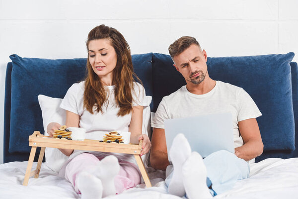 Husband using laptop and wife having breakfast in bed