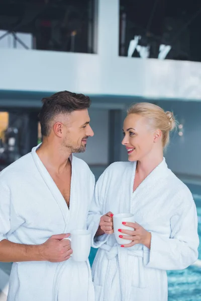 Pareja Sonriente Albornoces Pie Cerca Piscina Con Tazas — Foto de stock gratis