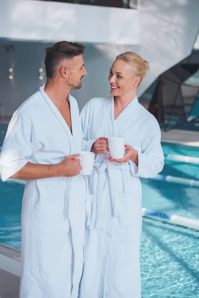 スイミング プールの近くに立って カップを保持している白いバスローブのカップルの笑顔 — ストック写真