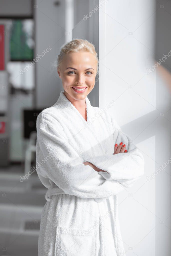attractive smiling adult woman in white bathrobe with arms crossed standing near window