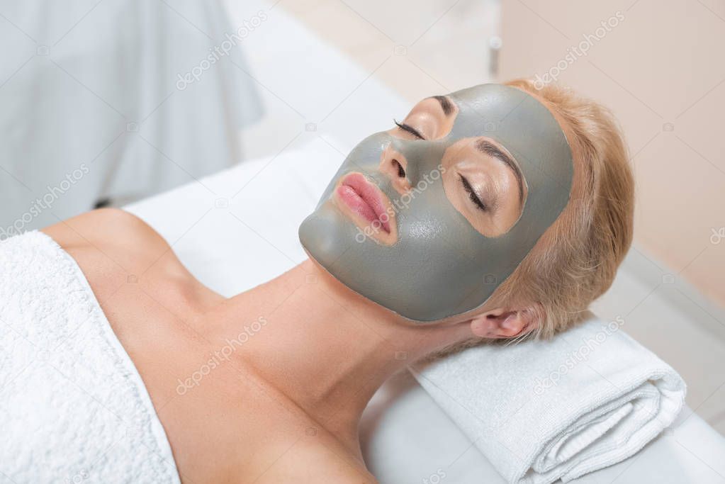 close up of blonde woman lying with facial clay mask in spa 