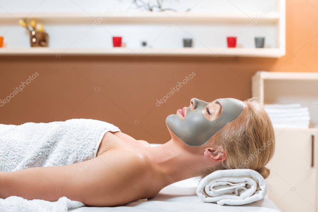 blonde woman lying on table with clay mask on face in spa