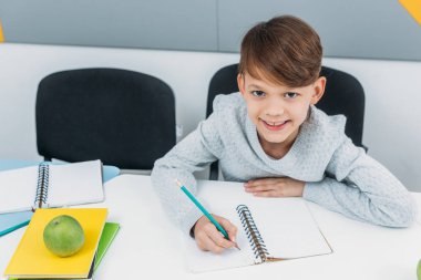 happy schoolboy writing in notepad in classroom clipart
