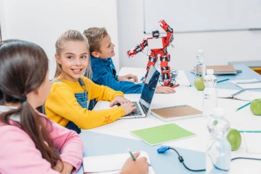 Not defterinde yazma ve kök sınıfta kırmızı robot ile masada dizüstü kullanarak çocuk