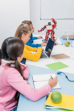 not defterlerinde yazma, dizüstü bilgisayar kullanarak ve kök sınıfta masada kırmızı robot bakarak çocuk