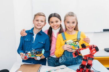happy schoolchildren looking at camera and holding mechanical robots in stem class clipart