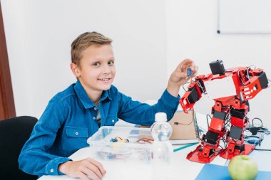 Kök sınıf robot modeli ile masada oturan ve kameraya bakarak okul çocuğu