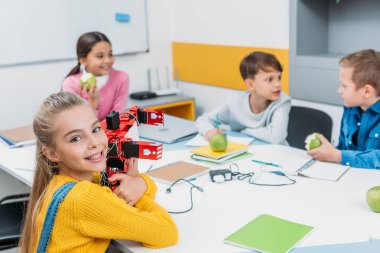 happy schoolgirl holding robot model while classmates eating apples on STEM project in classrom clipart