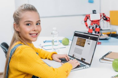 robot modeli ile masada oturan, kameraya bakıyor ve kök ders sırasında ekranda bilim sitesi ile dizüstü bilgisayar kullanarak kız öğrenci gülümseyen  