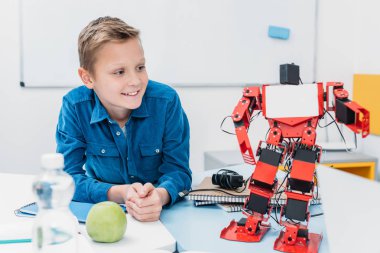 smiling schoolboy sitting at table and looking at robot model during STEM lesson   clipart