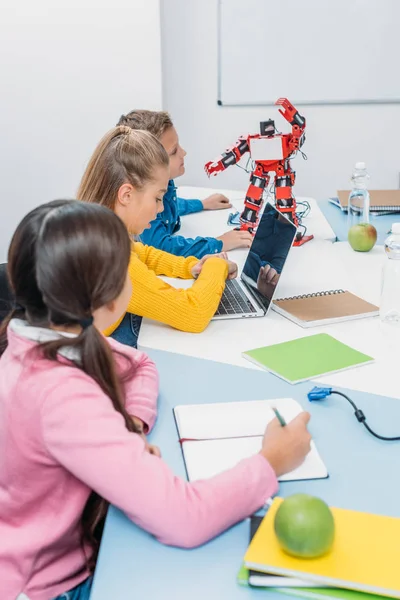 children writing in notebooks, using laptop and looking at red robot at desk in stem class