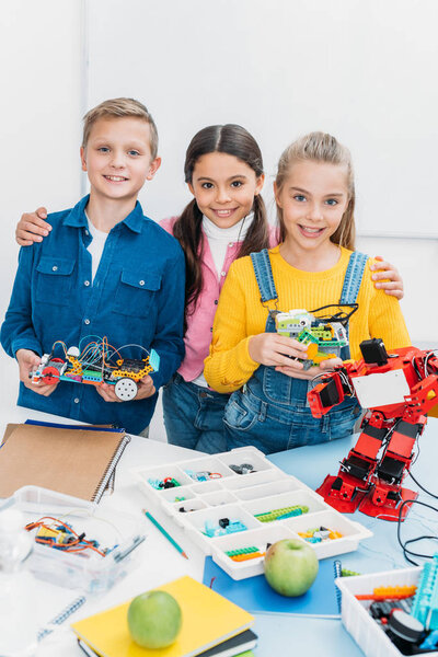 smiling schoolchildren looking at camera and holding handmade robots in classroom
